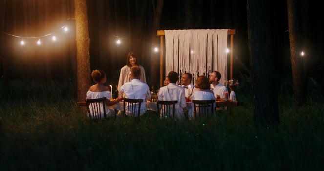 Guests smile sitting at a beautifully served festive table outdoors at night under the light bulbs, a woman stands saying congratulations photo