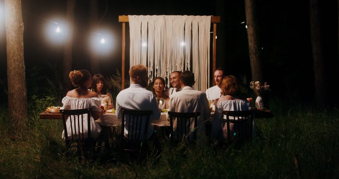 The bride and groom embrace sitting at the festive table next to their guests. A man takes pictures of the bride and groom on his mobile. Outdoor wedding feast photo