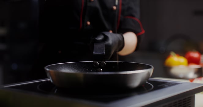 A woman in a chef's uniform with disposable black gloves on her hands pours oil on a frying pan standing on the stove, a close photo