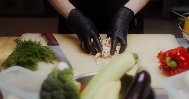A woman in a chef's uniform with disposable black gloves on her hands shifts chopped mushrooms from a cutting board into a frying pan, close photo