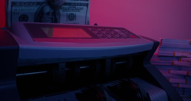 A man's hand with a ring inserts a stack of hundred-dollar bills into the counting machine and it starts counting. Bundles of money are lying around on the table - machine photo