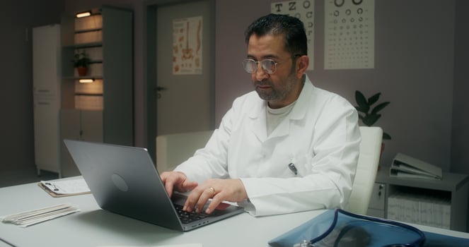 A female patient enters a medical office for a medical appointment. The doctor uses a laptop sitting at the table - enters photo