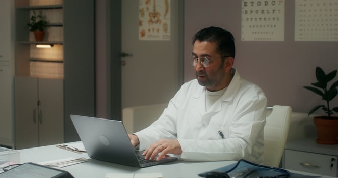 An elderly doctor looks at the camera with a serious look sitting in front of an open laptop in the medical office at the clinic - serious photo