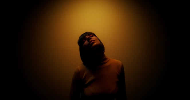 A woman in a wig dances moving her arms while standing in the orange color of a lamp in the studio photo