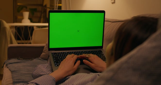 A woman typing on a laptop with a green screen lying on the couch in an apartment with dim lights, close-up of the screen, an unrecognizable person photo