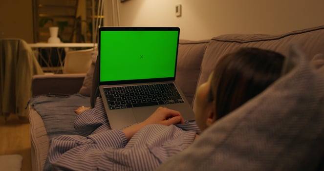 A woman uses a laptop with green screen lying on the couch in an apartment with dim lights photo