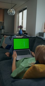 A woman uses a laptop with green screen lying on the couch at home, an unrecognizable person photo