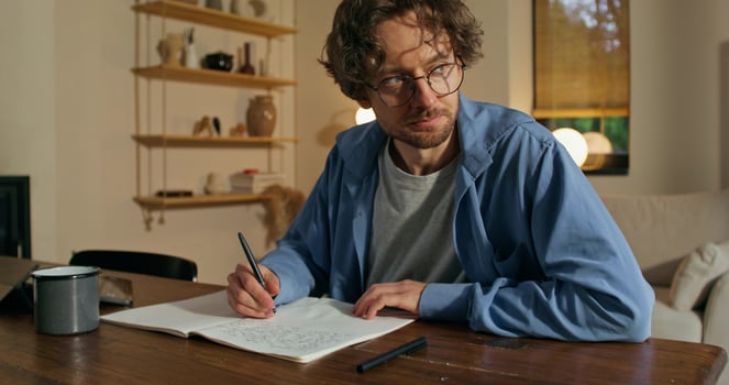 A man with glasses is writing in a notebook sitting at a table with a serious face. Stylish home interior - sitting photo