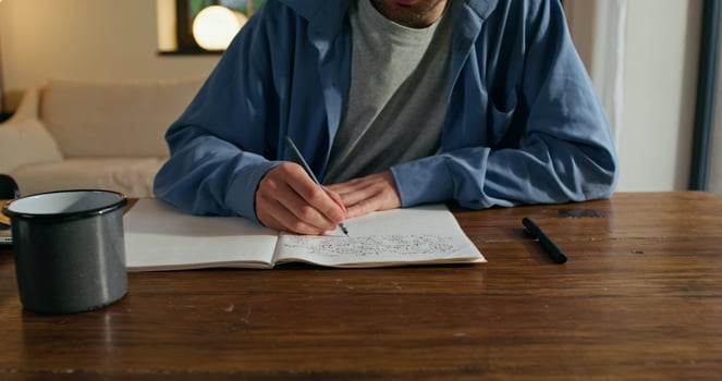 A man with glasses is writing in a notebook sitting at a table with a serious face. Stylish home interior photo