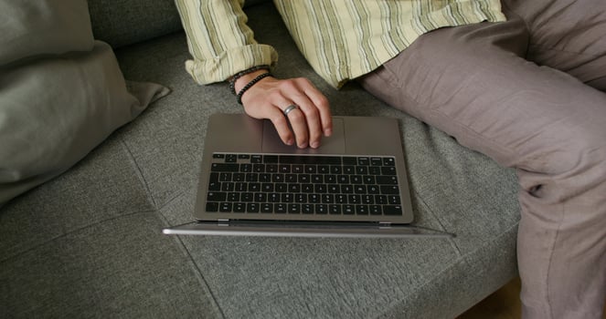 A bearded man uses a laptop and talks on a mobile phone sitting on the couch at home with a serious look. Daylight - mobile photo