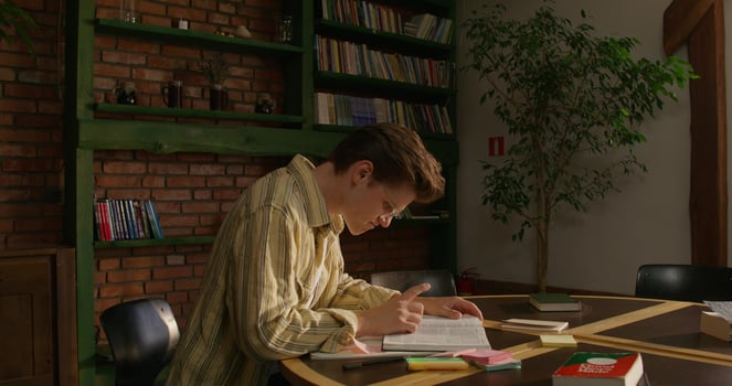 A man is studying the Bible while sitting at a table indoors. He prays with his hands folded in front of him. Daylight photo