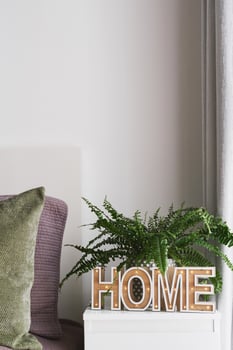 Bedroom interior, bedside table with 'Home' sign, potted fern plant and bed with green and purple pillows. Cozy home concept photo