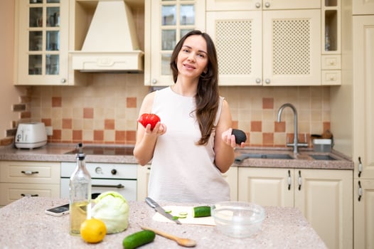 young woman in bright kitchen holding ripe tomato and avocado in hands and going to prepare salad. fresh vegetables and cutting board are on table photo
