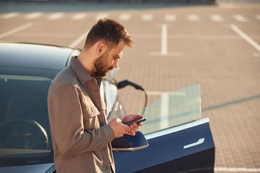 Bored,, waiting for automobile to charge... With smartphone. Man is standing near his electric car outdoors photo