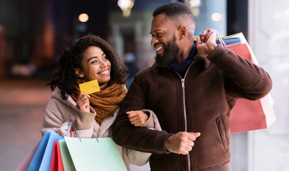 Let's Buy Something. Portrait of smiling black woman showing debit credit card to man, holding shopping bags, walking outside in the city near mall in the evening, blurred background photo