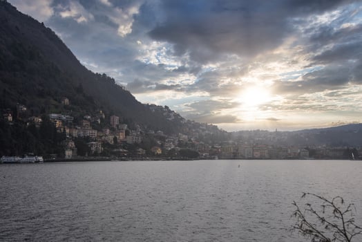 Lake Como at sunset with a forested hillside, colorful buildings, a docked ferry, and branches in the foreground under a partially cloudy sky. photo