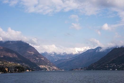 Lake Como's calm waters reflect the sky, surrounded by snow capped mountains, scattered towns, greenery, and rugged terrain along the shoreline. photo