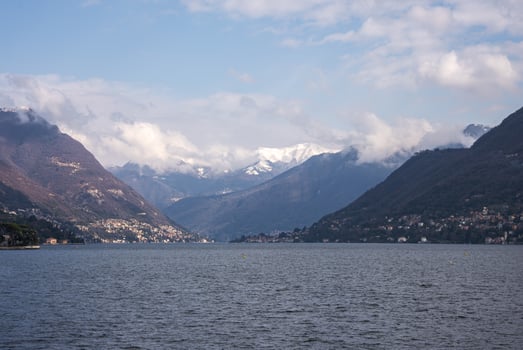 Lake Como is surrounded by forested hills and distant snow capped mountains. Small towns line the shoreline under a partly cloudy sky. photo