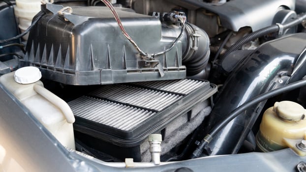 Vehicle engine compartment change equipment activity. Focus is on used rectangular air filter positioned within a black plastic housing. Various other engine components including hoses and wires. photo