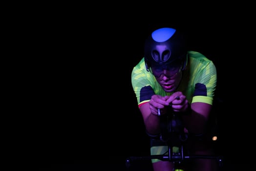 A triathlete rides his bike in the darkness of night, pushing himself to prepare for a marathon. The contrast between the darkness and the light of his bike creates a sense of drama and highlights the photo