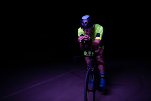 A triathlete rides his bike in the darkness of night, pushing himself to prepare for a marathon. The contrast between the darkness and the light of his bike creates a sense of drama and highlights the photo
