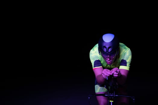 A triathlete rides his bike in the darkness of night, pushing himself to prepare for a marathon. The contrast between the darkness and the light of his bike creates a sense of drama and highlights the photo