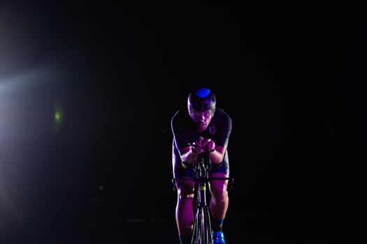 A triathlete rides his bike in the darkness of night, pushing himself to prepare for a marathon. The contrast between the darkness and the light of his bike creates a sense of drama and highlights the photo