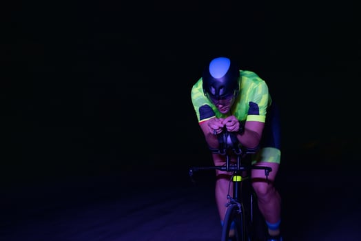 A triathlete rides his bike in the darkness of night, pushing himself to prepare for a marathon. The contrast between the darkness and the light of his bike creates a sense of drama and highlights the photo