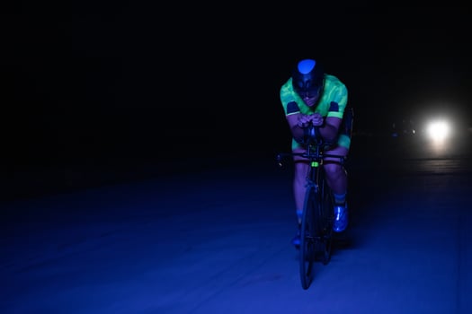 A triathlete rides his bike in the darkness of night, pushing himself to prepare for a marathon. The contrast between the darkness and the light of his bike creates a sense of drama and highlights the photo