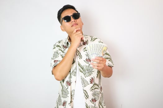 portrait of smiling asian man holding and showing money. portrait of indonesian man in hawaiian shirt looking at camera on isolated white background. gesture showing or thinking photo