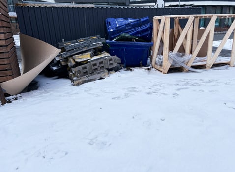 Construction debris stacked beside a black metal fence on a snowy urban street. Includes blue dumpsters, concrete slabs, wooden frames, and discarded materials photo