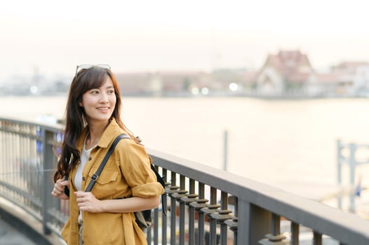 cheerful Asian woman with backpack enjoys serene riverside view during sunset, standing by railing with soft smile photo