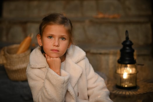 The girl sits quietly on the steps, a warm lamp glowing behind her. Beside her, a vintage-style basket with fresh baguettes adds a cozy, timeless touch. photo