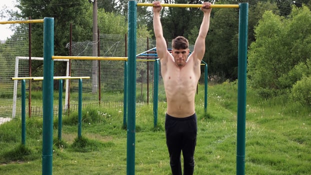 Muscular athlete doing pull ups on horizontal bar at sports ground. Young shirtless sportsman exercising at nature. Strong sporty man training outdoor. Concept of healthy and active lifestyle. Slow photo