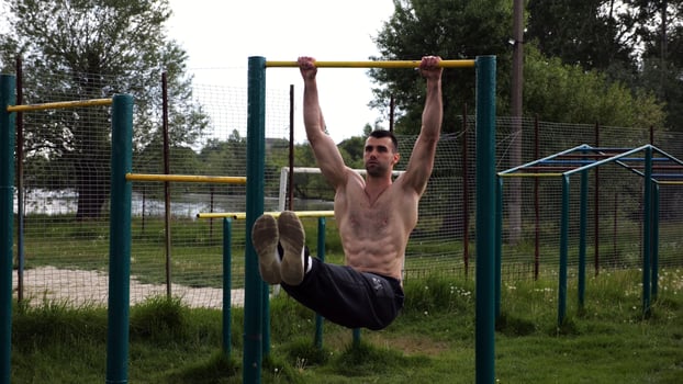 Athletic man trains abdominal muscles raising legs on horizontal bar at sports ground. Young sportsman exercising on pull-up bar outdoor. Sporty athlete working out at nature. photo