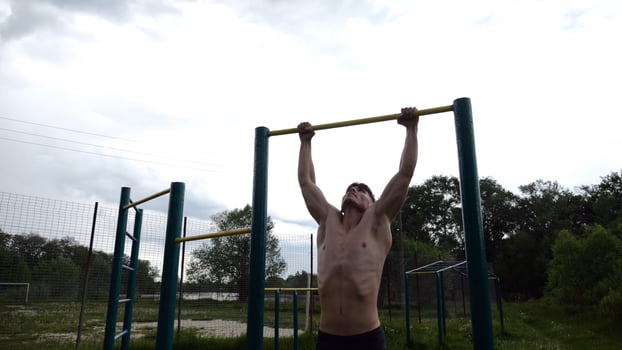 Muscular athlete doing pull ups on horizontal bar at sports ground. Young shirtless sportsman exercising at nature. Strong sporty man training outdoor. Concept of healthy and active lifestyle. photo