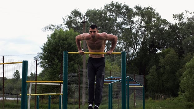 Hardy shirtless sportsman doing push ups on horizontal bar at nature. Strong and muscular guy training at sports ground. Young athletic guy exercising outdoor. Concept of active and healthy lifestyl photo