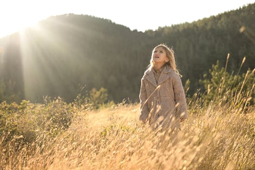 In the soft glow of sunrise, a little girl with light hair and a pink coat gazes peacefully across a field of tall golden grass photo
