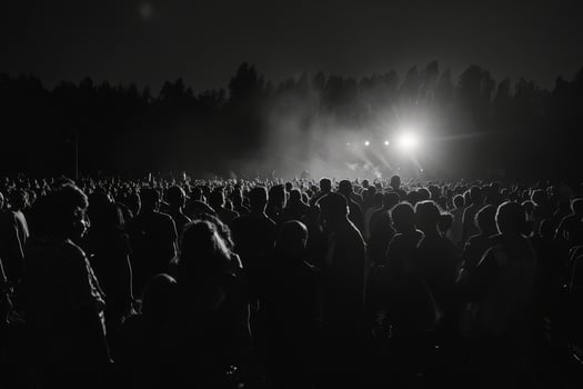A large crowd stands silhouetted against bright stage lights, enjoying a live performance in an outdoor setting during nighttime photo