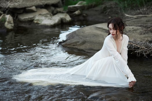 Erotic young woman in a long white dress walks in spring park near river. Sexy brunette posing in nature photo