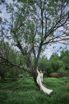Erotic young woman in long white dress walks in spring park near river. Sexy brunette posing in nature photo