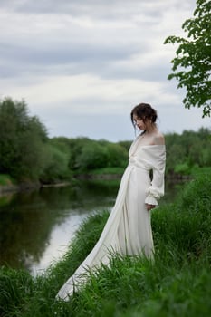 Beautiful young woman in white dress resting on shore lake pond river. Professional makeup and hairstyle with curled hair. photo