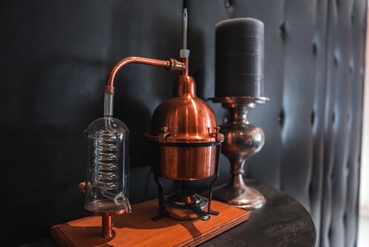 A vintage copper distillation apparatus with a glass condenser sits on a wooden base beside a black candle in a silver holder on tufted black leather. photo