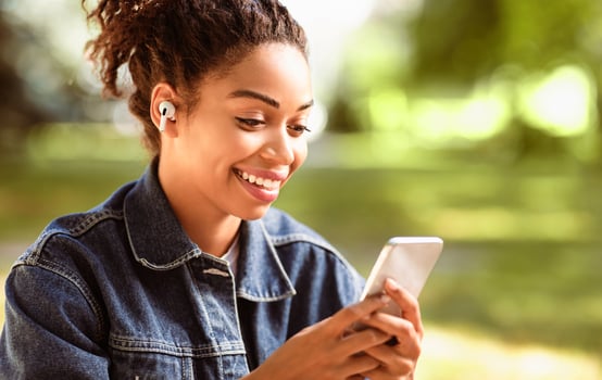 Cheerful Black Woman Using Mobile Phone And Earbuds Earphones Siitting In Park Outdoor. Music Application Concept photo