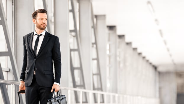 Middle-aged handsome businessman walking by airport terminal with luggage and case, looking at copy space, panorama. Rich entrepreneur in black suit going by airport, departuring for business trip photo
