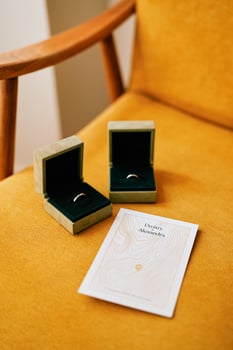 Wedding rings in boxes lie in a chair next to a personalized invitation photo