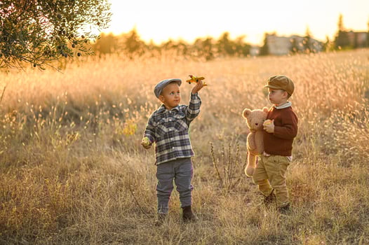 Dressed in retro clothing, two boys enjoy a sunset adventure in the field with a toy plane, an apple, and a stuffed bear. photo
