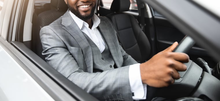 Smiling young black businessman in stylish suit driving luxury car, looking through car window and smiling at camera, holding hands on wheel, going to office for business meeting photo
