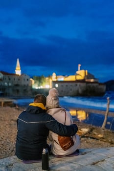 Man and woman sit embracing on the seashore and look at the illuminated old houses. Back view photo