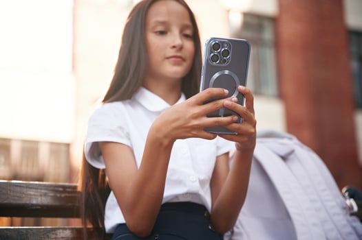 Silver, colored smartphone in hands. School girl in uniform is outdoors near the building photo